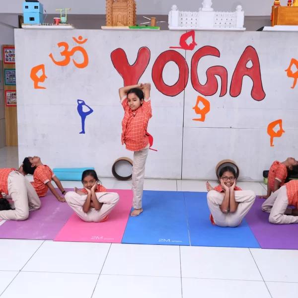Children practicing yoga on colorful mats in a classroom, with a ‘YOGA’ sign and decorations on the wall behind them.