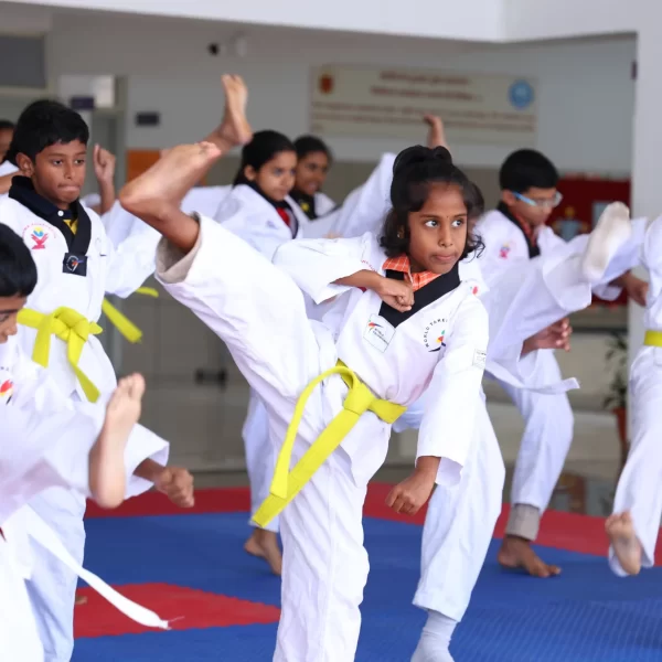 Group of children practicing martial arts in a dojo, wearing white uniforms with yellow belts and performing high kicks under an instructor’s supervision.