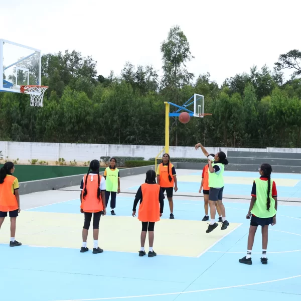 Group of children playing basketball on an outdoor court, with one child shooting the ball while others in colored jerseys watch and defend.