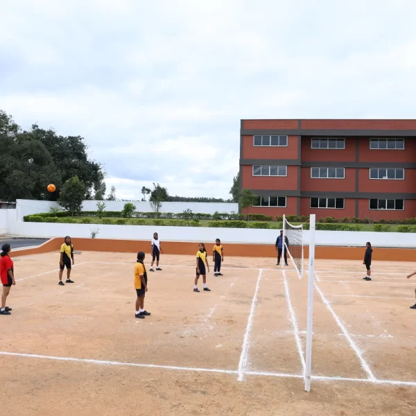 A group of students playing volleyball on an outdoor court with clearly marked white boundary lines, set in front of a red multi-story school building under a cloudy sky.