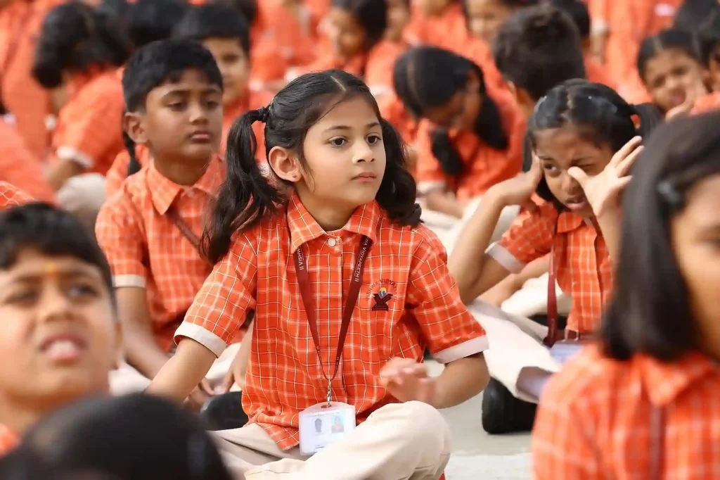 Students in school uniforms are seated outdoors, with a young girl in the center looking attentive amidst a group of peers.