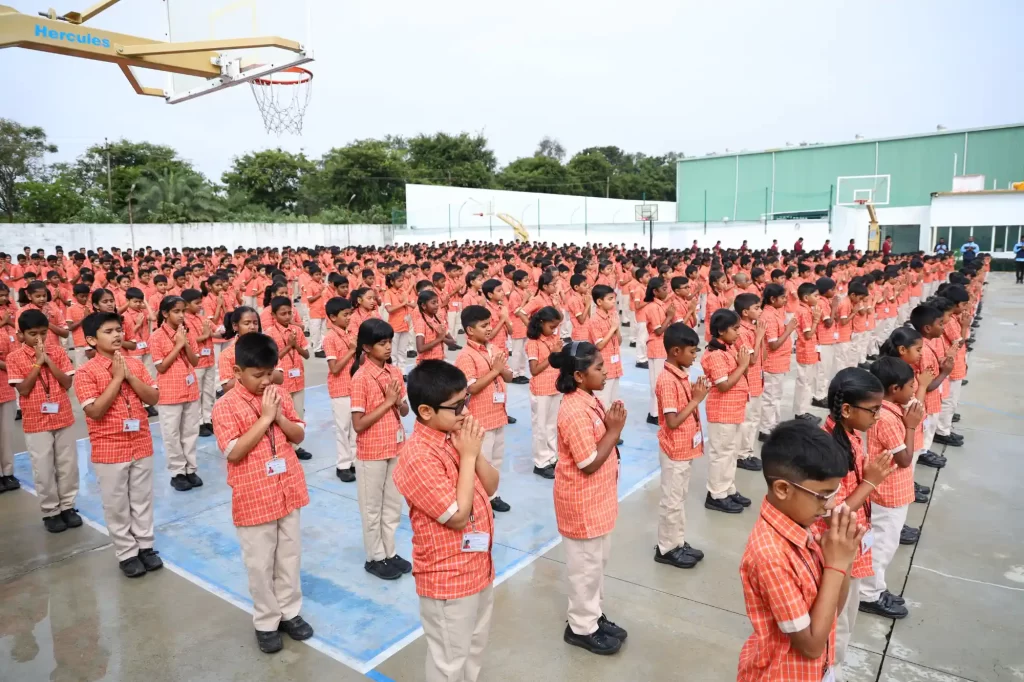 Students in orange uniforms are standing in rows, participating in an outdoor assembly on a sports court.