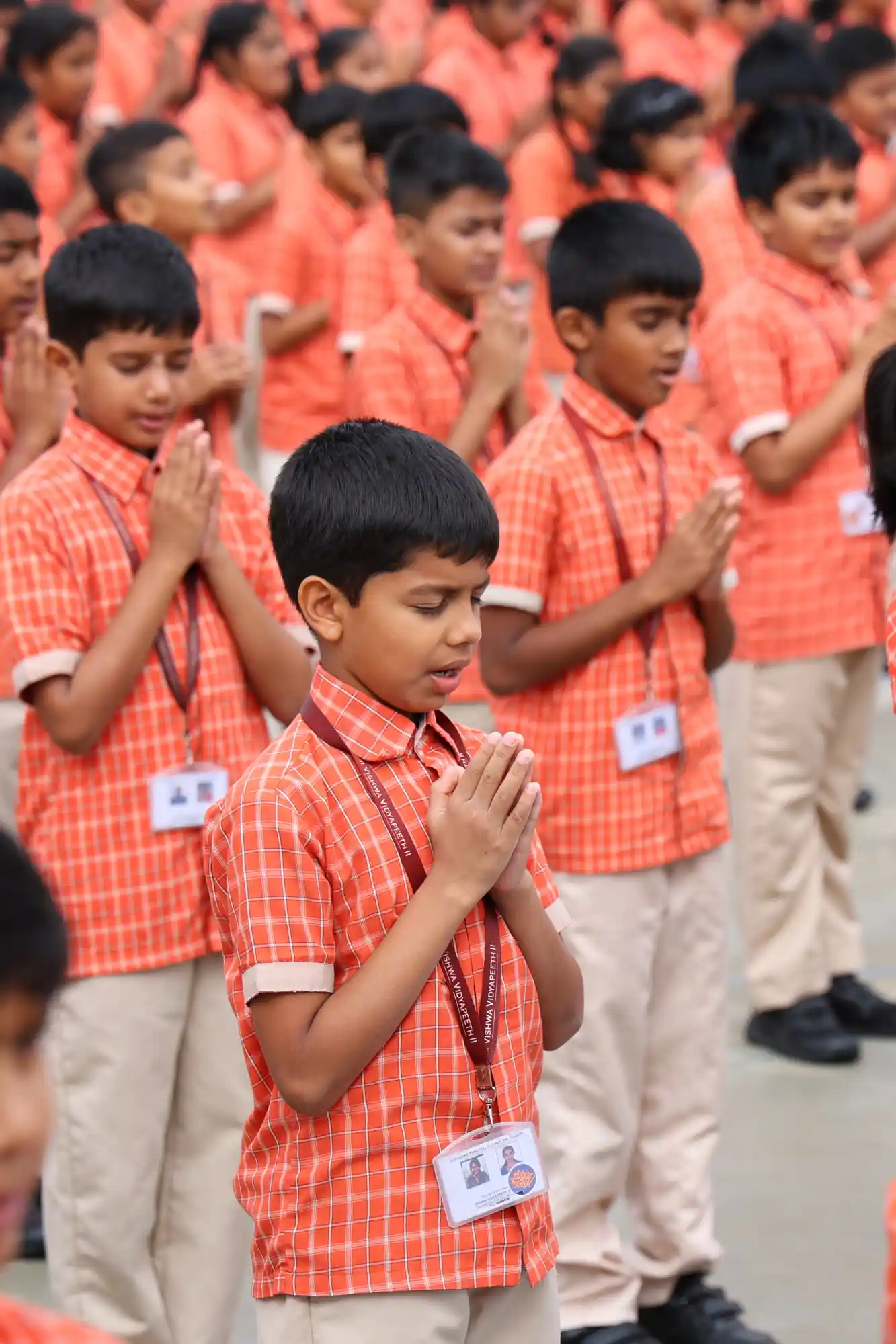 Group of school children dressed in orange uniforms, standing in a prayer pose during a school assembly