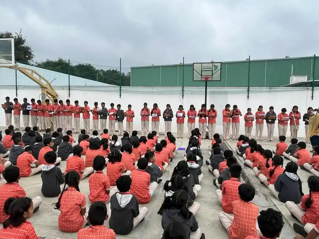 Students gathered in an outdoor assembly, sitting in neat rows, with a line of teachers or students standing in front of a basketball hoop in the background.