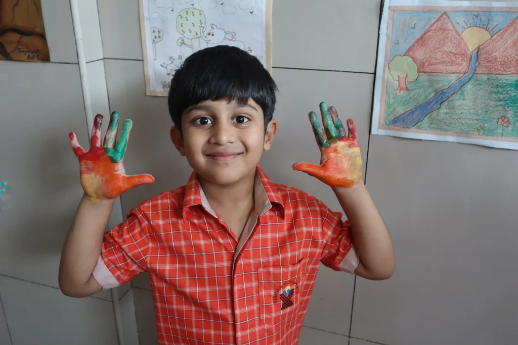 A smiling child in an orange uniform shows painted hands in a classroom setting with artwork on the wall.