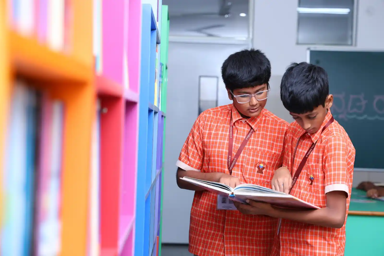 Two students in orange checkered uniforms are standing near a bookshelf, focused on reading a large open book together.