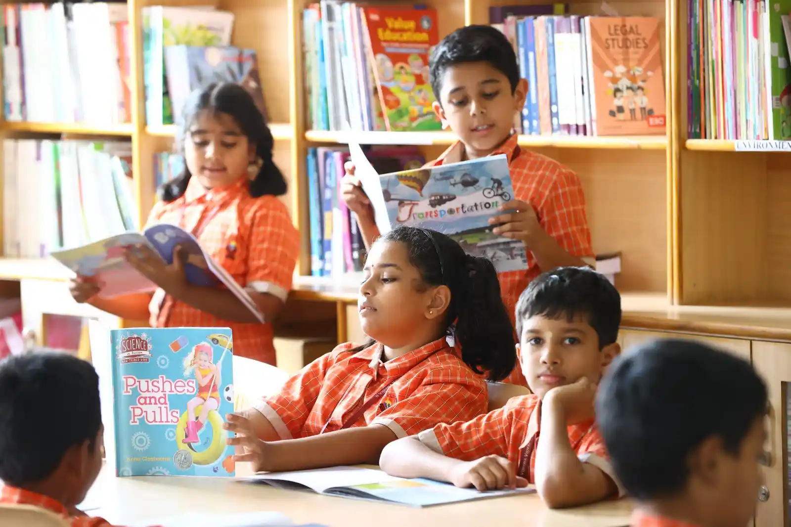 Children are engaged in reading books in a library, surrounded by shelves filled with colorful books.







