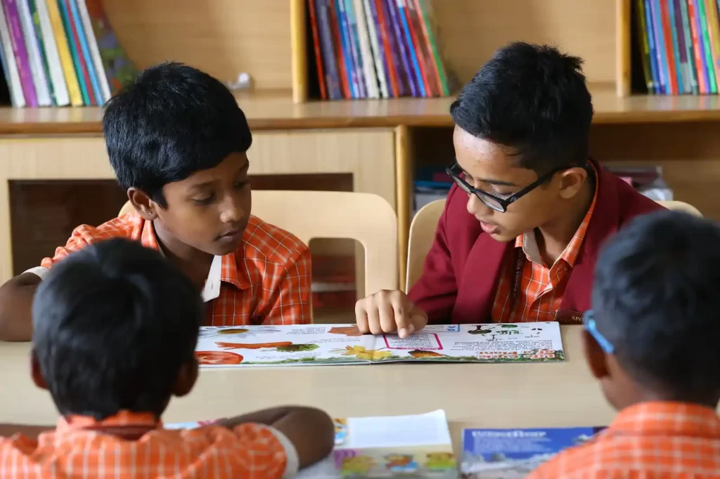 Children are seated around a table in a library, collaboratively reading and discussing a book, with bookshelves in the background.