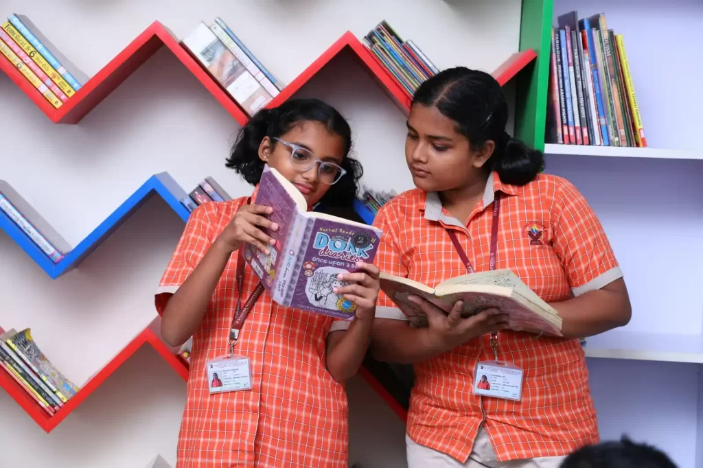 Two students in school uniforms are reading books in front of a colorful zigzag bookshelf filled with books.