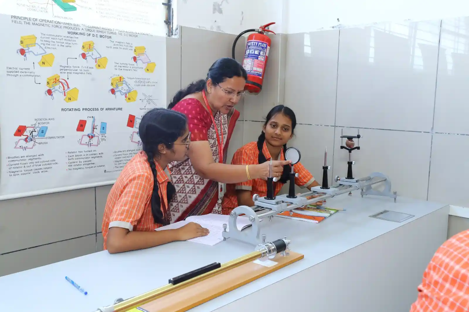 A teacher assists two students in a lab setting with measurement equipment, a wall chart, and a fire extinguisher in the background.