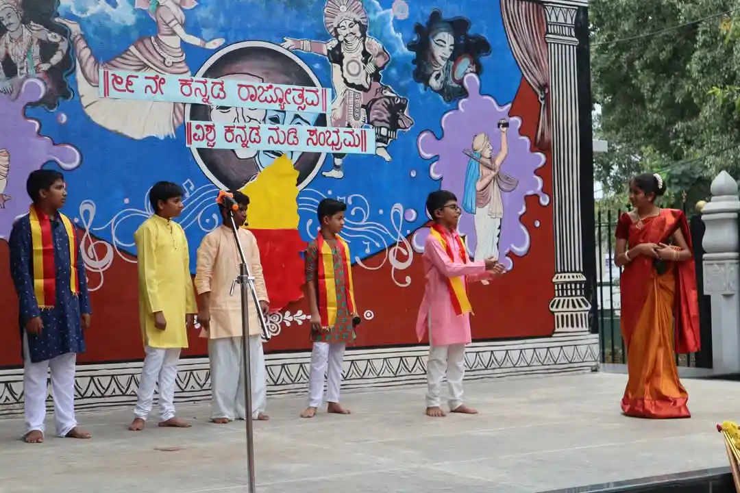 A group of students is performing on an outdoor stage, dressed in traditional attire. A decorated backdrop featuring artwork and text is behind them, and one student appears to be addressing the audience with a microphone.