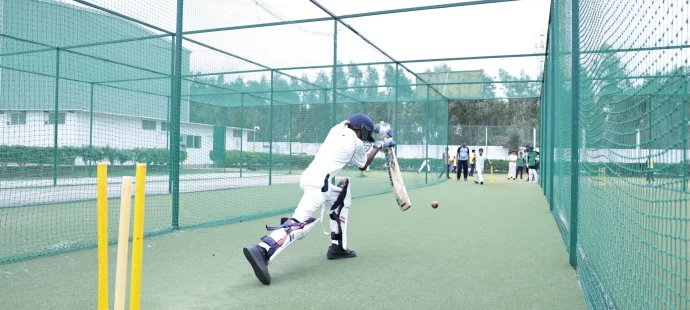 A cricketer practices batting in an outdoor net as others watch in the background.