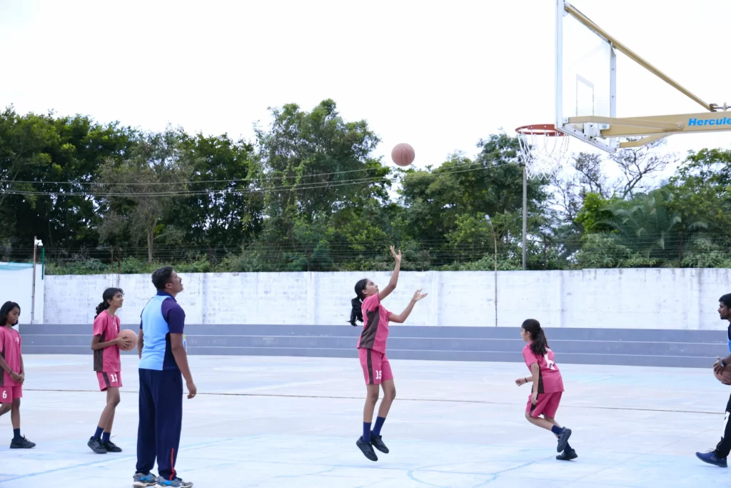 A girl shoots a basketball while others, in pink sportswear, watch. A coach stands nearby holding a ball, with trees and a wall in the background.