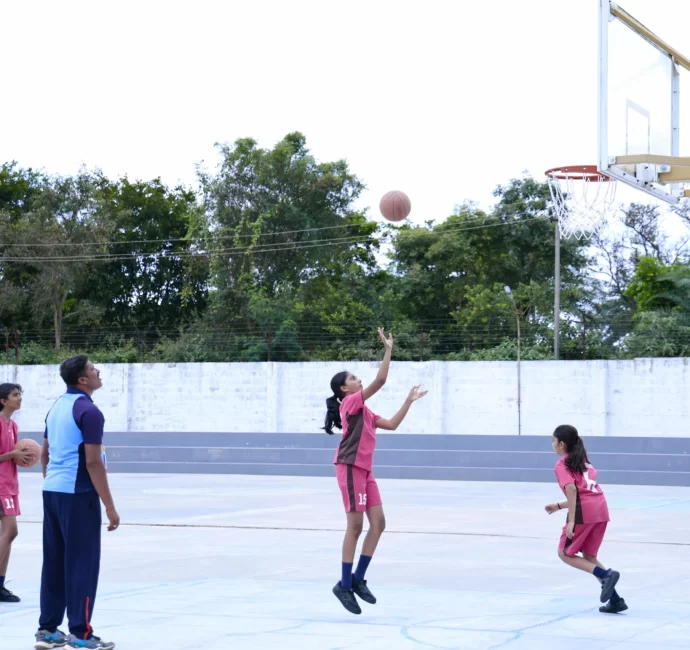 A girl shoots a basketball while others, in pink sportswear, watch. A coach stands nearby holding a ball, with trees and a wall in the background.