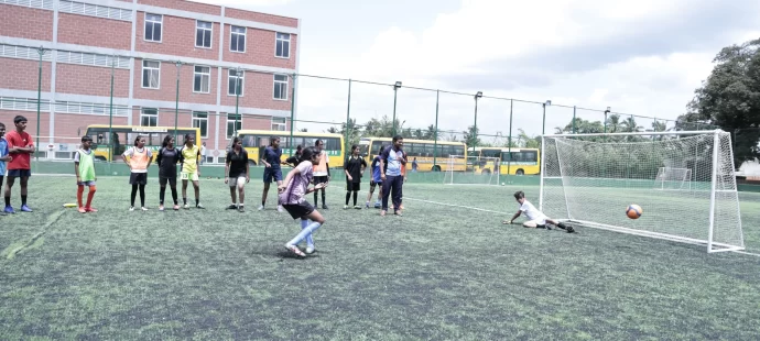 A soccer player scores a penalty while a group watches. The goalkeeper dives, but the ball is already in the net. School buses and a building are in the background.