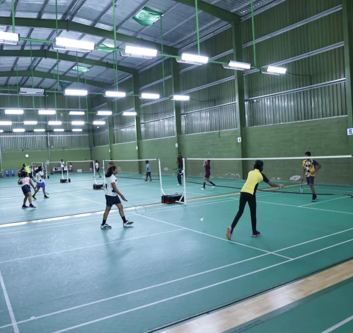 Students play badminton in a large, well-lit indoor court with green flooring and walls.