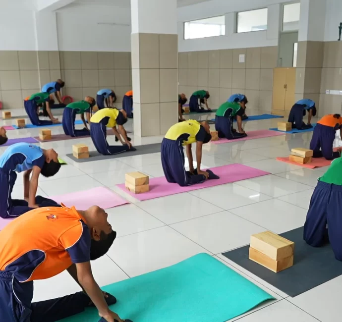 Students in a classroom practice yoga, performing back-bending poses on mats with yoga blocks, dressed in colorful uniforms.