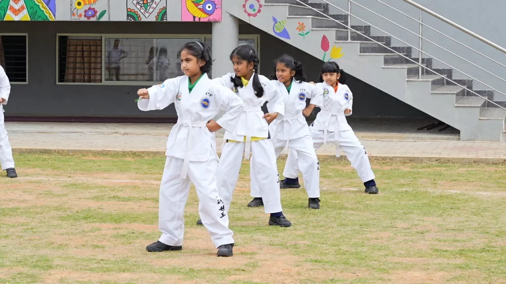 Young girls in martial arts uniforms practice in an outdoor area, performing synchronized movements.