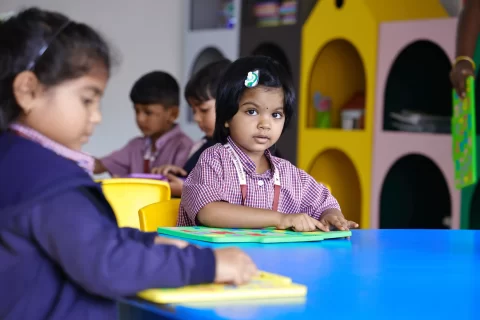 Young children in school uniforms seated at a blue table in a classroom, engaging with learning materials, with one girl looking toward the camera.