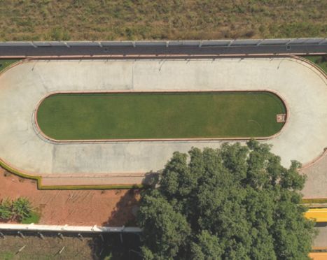 Aerial view of an oval sports track with a grass field in the center, surrounded by trees and a boundary wall.