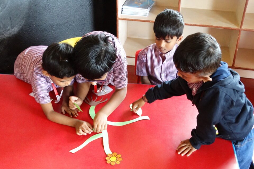 Children gathered around a red table, working together on assembling a plant craft, with one child holding a piece of the plant stem.