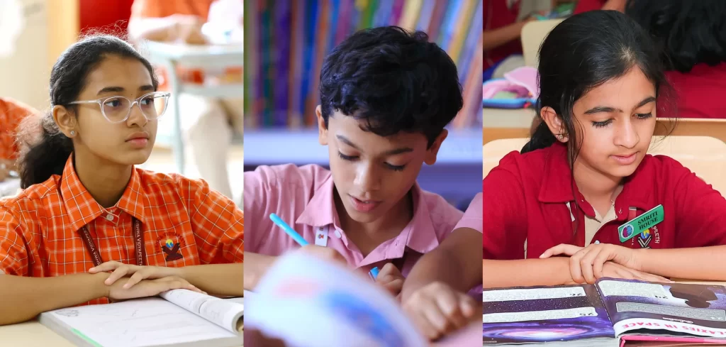 Three school-aged children sitting at desks in a classroom, focused on reading or writing. The child on the left wears glasses and an orange uniform, the middle child is reading a book, and the child on the right is in a red uniform, also reading attentively.