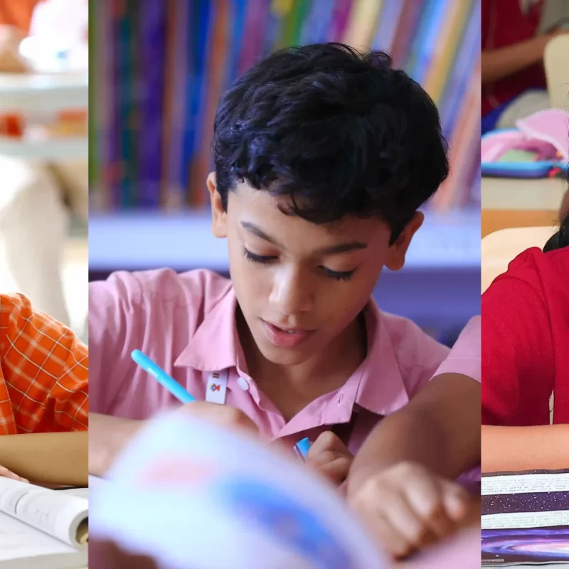 Three school-aged children sitting at desks in a classroom, focused on reading or writing. The child on the left wears glasses and an orange uniform, the middle child is reading a book, and the child on the right is in a red uniform, also reading attentively.